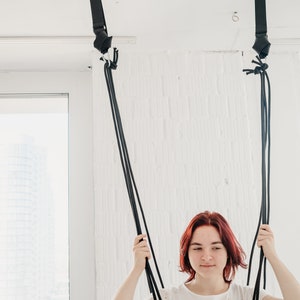 May include: A woman with red hair is sitting in a black and blue fabric swing hanging from the ceiling. The swing is made of two straps that are attached to the ceiling with hooks.