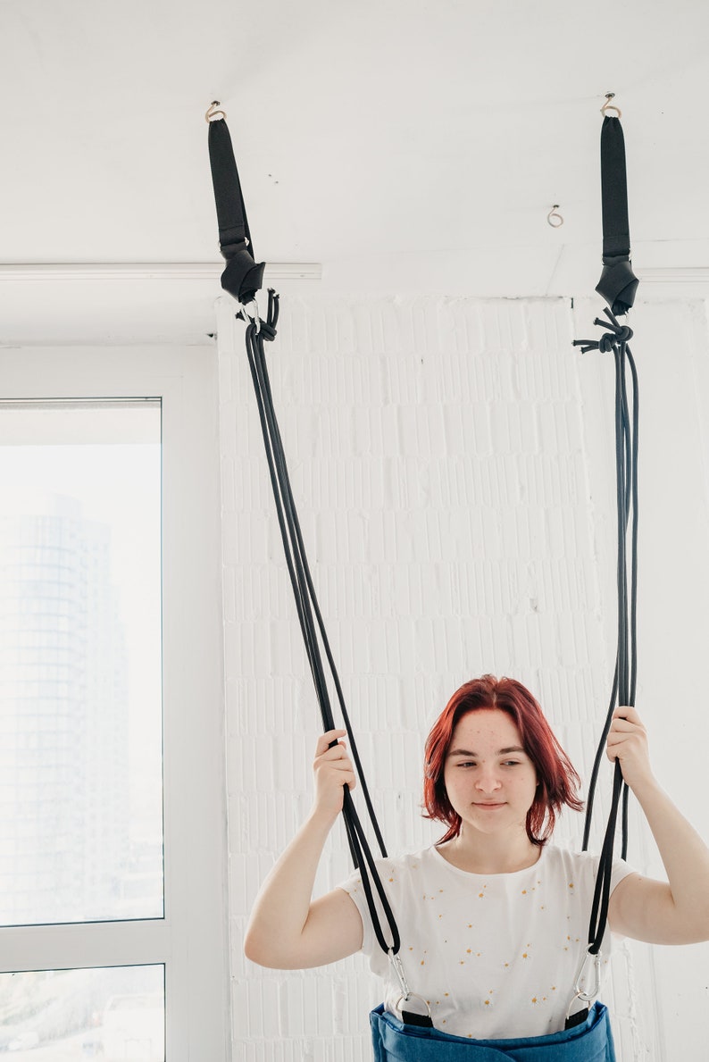 May include: A woman with red hair is sitting in a black and blue fabric swing hanging from the ceiling. The swing is made of two straps that are attached to the ceiling with hooks.