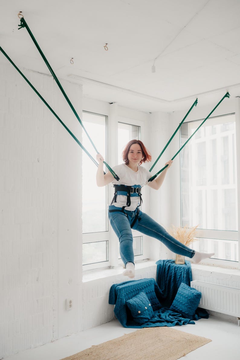 Pode incluir: Um sistema de arn&ecirc;s de suspens&atilde;o azul e preto com al&ccedil;as ajust&aacute;veis e suporte de cintura acolchoado, conectado a quatro cordas verde-escuras ancoradas ao teto, projetado para fitness a&eacute;reo ou atividades de movimento terap&ecirc;utico.