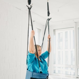 May include: A person wearing a blue shirt and jeans is suspended from the ceiling by a blue and black harness. The harness is attached to two black straps that are connected to hooks in the ceiling.