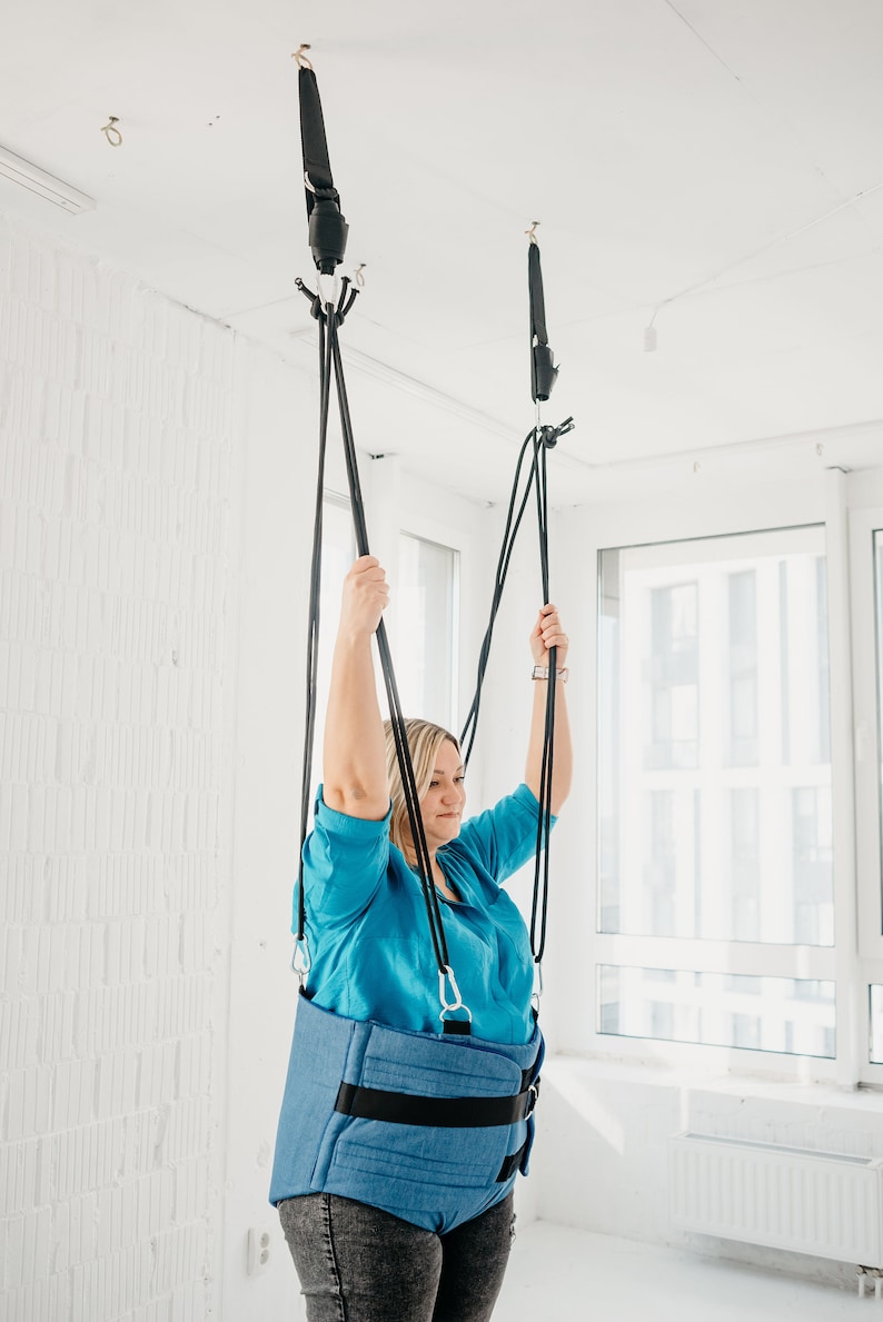May include: A person wearing a blue shirt and jeans is suspended from the ceiling by a blue and black harness. The harness is attached to two black straps that are connected to hooks in the ceiling.