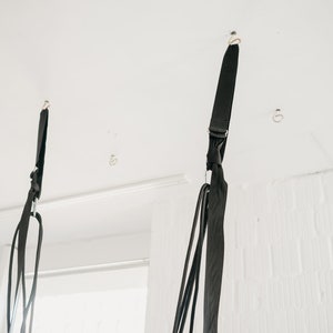 May include: Two black straps hanging from the ceiling, each attached to a hook. The straps are made of a woven material and have a silver metal ring at the bottom.