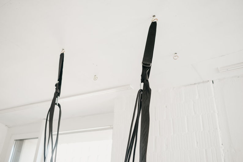 May include: Two black straps hanging from the ceiling, each attached to a hook. The straps are made of a woven material and have a silver metal ring at the bottom.