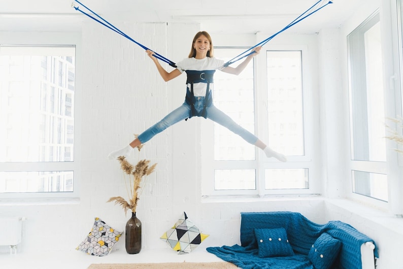 May include: A young person wearing a white shirt and blue jeans is suspended in mid-air by a blue bungee cord system. They are smiling and looking at the camera. The person is in a white room with windows and a blue couch in the background.
