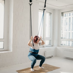 May include: A woman wearing a blue harness is doing a squat exercise while suspended from the ceiling by two black straps.