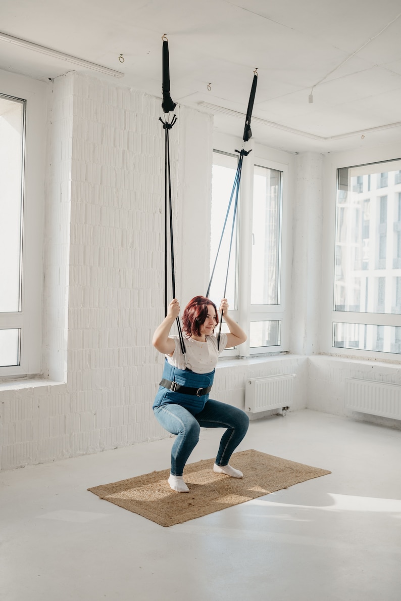May include: A woman wearing a blue harness is doing a squat exercise while suspended from the ceiling by two black straps.