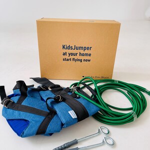 Pode incluir: Um conjunto KidsJumper com um arn&ecirc;s de tecido azul com tiras pretas, uma corda el&aacute;stica longa verde e dois parafusos de olhal met&aacute;licos com buchas de pl&aacute;stico. Os itens est&atilde;o em frente a uma caixa de papel&atilde;o com o texto KidsJumper at your home start flying now.