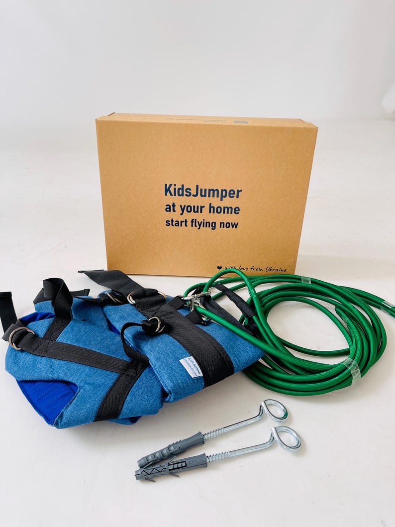 Pode incluir: Um conjunto KidsJumper com um arn&ecirc;s de tecido azul com tiras pretas, uma corda el&aacute;stica longa verde e dois parafusos de olhal met&aacute;licos com buchas de pl&aacute;stico. Os itens est&atilde;o em frente a uma caixa de papel&atilde;o com o texto KidsJumper at your home start flying now.