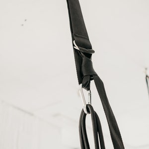 May include: Black fabric straps with a silver carabiner hook hanging from a white ceiling. The straps are tied together in a knot.