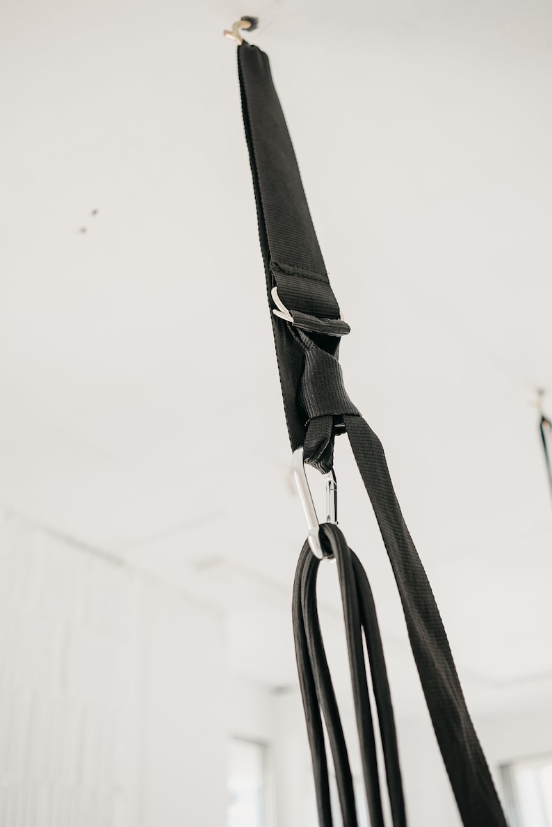 May include: Black fabric straps with a silver carabiner hook hanging from a white ceiling. The straps are tied together in a knot.