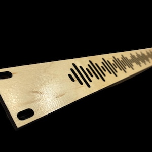 May include: A light-colored wooden sound wave art piece. The piece is rectangular with a series of cut-out lines forming a sound wave pattern. Two oval holes are cut out near the base. The piece is angled, creating a shadow.
