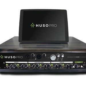 May include: A HUSO PRO audio device with a dark wood finish and a black control panel featuring multiple knobs and inputs. A tablet is mounted on top displaying the HUSO PRO logo. The panel includes labels for WRISTS, FLOATING PADS, and ANKLES.