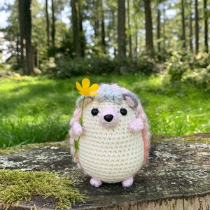 Puede incluir: Un juguete de erizo de crochet que sostiene una flor amarilla. El erizo es blanco con detalles rosas y grises. El juguete está sentado en un tocón de árbol cubierto de musgo.