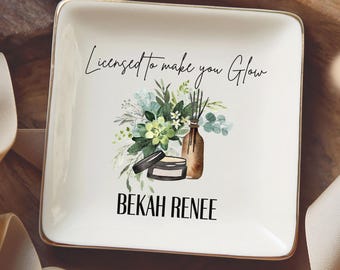 May include: A square white ceramic trinket dish with a gold-toned rim. The center features a graphic of a brown bottle, a small jar, and green foliage. The text reads Licensed to make you Glow and the name BEKAH RENEE in black lettering.