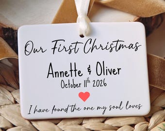 May include: A rectangular white ceramic ornament hanging from a white ribbon. The text reads Our First Christmas, Annette & Oliver, October 11th 2026, with a small red heart and the phrase I have found the one my soul loves printed in black.