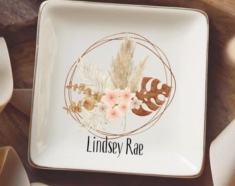 May include: A square white ceramic trinket dish with a gold-toned rim. The center features a circular line drawing enclosing a botanical arrangement of pampas grass, leaves, and small flowers, with the name Lindsey Rae printed below.