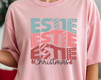 May include: A light pink short-sleeve t-shirt featuring a graphic print with the word ESTIE repeated three times in teal, light pink, and red, followed by the word Christmas in cursive script with small red tree icons.