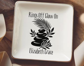 May include: A square white ceramic trinket dish with a gold-toned rim. The center features a black graphic of stacked stones and palm leaves with the text "Rings Off Glow On" above and the name "Elizabeth Grace" printed below the design.