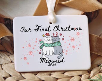 May include: A rectangular white ceramic ornament hanging from a white ribbon. The front features the text Our First Christmas and Meowied 2026 with an illustration of two cats wearing a green scarf, a Santa hat, and reindeer antlers.