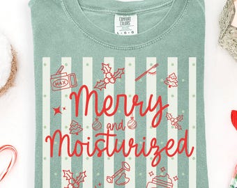 May include: A sage green t-shirt featuring a white vertical striped pattern and red graphic icons of holly, ornaments, and skincare jars. The text reads Merry and Moisturized in a red script font. The shirt has a crew neckline and short sleeves.