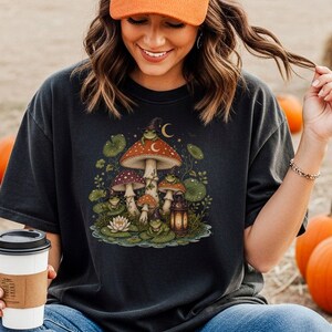Puede incluir: Una camiseta negra con una ilustración gráfica de varias setas, nenúfares y pequeñas ranas. Una rana lleva un sombrero de bruja y una pequeña linterna se encuentra cerca de la base de las setas. El diseño incluye una luna creciente y estrellas.
