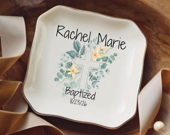 May include: A square white ceramic trinket dish with scalloped edges, featuring a printed design of a cross surrounded by eucalyptus leaves and yellow flowers, with the text Rachel Marie, Baptized, and the date 8/23/26 in black script.