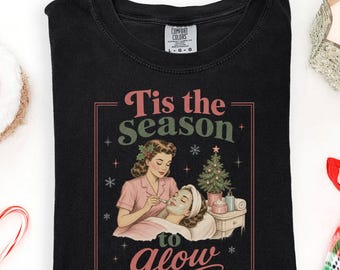 May include: A black t-shirt featuring a graphic of a person applying a facial mask to another person. The text reads 'Tis the season to glow'. The design includes a small Christmas tree, stars, and snowflakes in a rectangular frame.