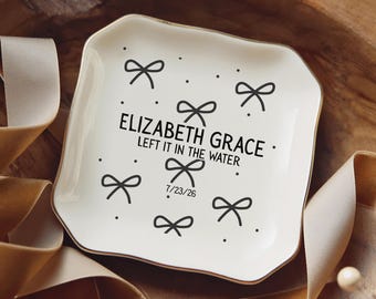 May include: A square white ceramic trinket dish with a gold-toned rim, featuring a black printed pattern of small bows and dots. The text reads ELIZABETH GRACE, LEFT IT IN THE WATER, and the date 7/23/26 in a simple black sans-serif font.