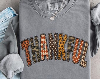 May include: A grey crewneck sweatshirt featuring the word THANKFUL in large, multi-patterned letters including leopard print, camouflage, polka dots, and diamond shapes, paired with light blue denim shorts with a frayed hem.