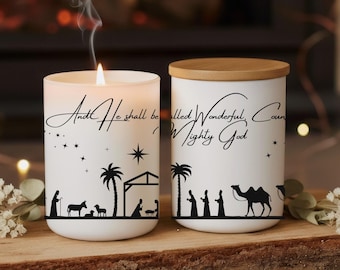 May include: Two white cylindrical candles featuring black silhouettes of a nativity scene. One candle is lit with a visible flame and smoke, while the other has a wooden lid. The text reads: And He shall be called Wonderful, Counselor, Mighty God.
