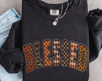 May include: A black crewneck sweatshirt featuring the word BLESSED in large, fabric-appliqued letters. Each letter displays a different pattern, including leopard print, camouflage, polka dots, and argyle. A silver chain necklace rests on the collar.