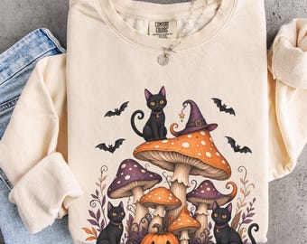 May include: A cream-colored long-sleeve sweatshirt featuring a graphic print of black cats, orange and purple mushrooms, a witch hat, and bats. The sweatshirt has a crew neckline and is paired with blue denim jeans. The word ivory is written on the side.