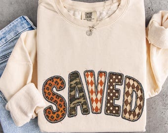 May include: A cream-colored long-sleeve sweatshirt featuring the word SAVED in large, fabric-appliqued letters. Each letter displays a different pattern, including leopard print, camouflage, orange diamonds, brown polka dots, and brown plaid.