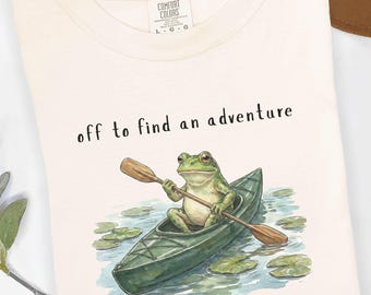 May include: An ivory t-shirt featuring a graphic of a green frog paddling a dark green kayak on water with lily pads. The text above the frog reads, "off to find an adventure." The shirt has a Comfort Colors brand label at the inner neckline.