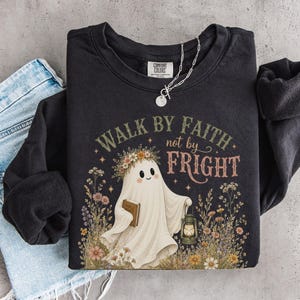 May include: A black crewneck sweatshirt featuring a graphic of a white ghost wearing a flower crown, holding a book and a lantern, surrounded by wildflowers. The text reads WALK BY FAITH not by FRIGHT. A silver chain necklace rests on the collar.