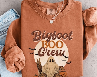 May include: A rust-colored crewneck sweatshirt featuring a graphic of a ghost wearing a Bigfoot costume, holding a pumpkin bucket, and making a peace sign. The text reads Bigfoot BOO Crew, surrounded by bats and a full moon design.