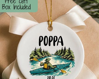May include: A round white ceramic ornament featuring a graphic of a person kayaking on a river surrounded by trees. The text POPPA is printed in black above the illustration, and the year 2026 is printed in black below the illustration.