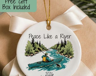 May include: A round white ceramic ornament featuring a graphic of a person in a teal kayak on a river with green trees. The text reads Peace Like a River, 2026, and Lindsey Rae. It hangs from a gold string against a gift box with a white ribbon.