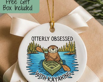 May include: A round white ceramic ornament featuring a graphic of an otter in a green kayak on blue water with pine trees in the background. The text reads OTTERLY OBSESSED WITH KAYAKING and 2026. The ornament hangs from a gold string.