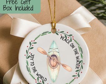 May include: A round white ceramic ornament featuring a central illustration of a kayak with a wooden paddle. A green pine wreath with red berries encircles the kayak. The text reads JOY TO THE WORLD and PEACE ON THE WATER.