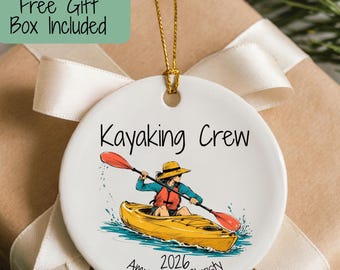 May include: A round white ceramic ornament featuring a colorful illustration of a person in a yellow kayak with a red paddle. The text reads Kayaking Crew, 2026, and the names Amy, Jorie, Christy. It hangs from a gold string.