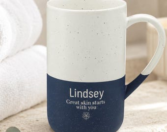 May include: A two-toned ceramic mug with a speckled white upper section and a solid navy blue lower section. The navy portion features the white text 'Lindsey', 'Great skin starts with you', and a small floral icon centered below.