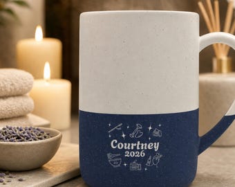 May include: A two-toned ceramic mug with a speckled white top and a speckled navy blue bottom. The navy section features white line art icons and the text "Courtney 2026" printed in the center. The mug has a curved handle on the right side.