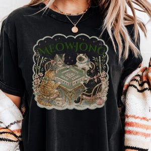 K&ouml;nnte beinhalten: Ein schwarzes T-Shirt mit einem Grafikdruck von vier Katzen, die an einem Tisch sitzen und Mahjong spielen. Der Text MEOWJONG ist in Gr&uuml;n &uuml;ber den Katzen gedruckt. Die Illustration enth&auml;lt Bambusstangen und florale Akzente in einem Rand.