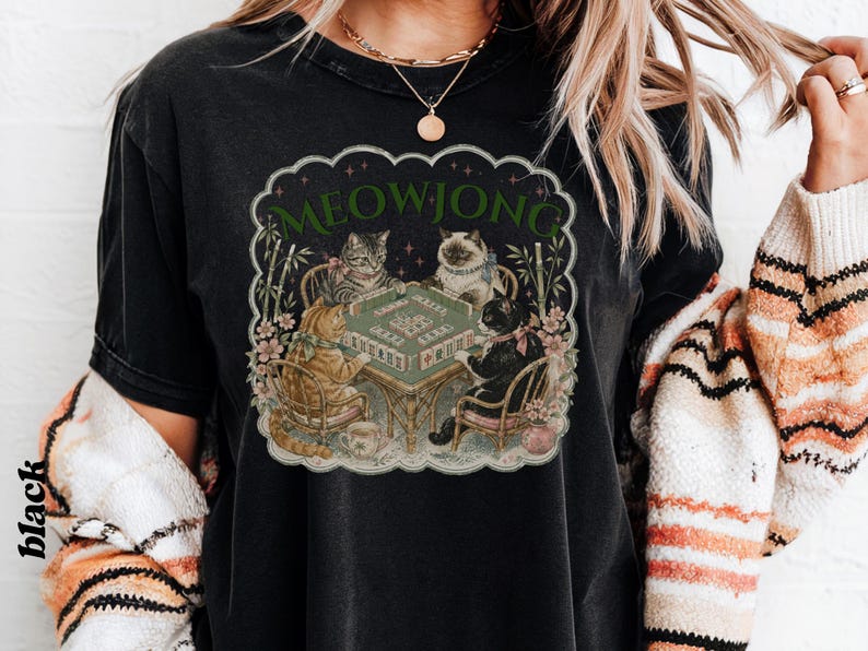 K&ouml;nnte beinhalten: Ein schwarzes T-Shirt mit einem Grafikdruck von vier Katzen, die an einem Tisch sitzen und Mahjong spielen. Der Text MEOWJONG ist in Gr&uuml;n &uuml;ber den Katzen gedruckt. Die Illustration enth&auml;lt Bambusstangen und florale Akzente in einem Rand.