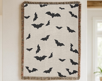 May include: A rectangular woven throw blanket featuring a repeating pattern of black bat silhouettes against a light beige background. The blanket has a textured fabric appearance and is finished with decorative fringed edges on all four sides.