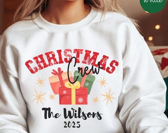 Könnte beinhalten: Weißes Sweatshirt mit weihnachtlichem Design. Der rote Text "CHRISTMAS Crew" steht über einer Grafik von verpackten Geschenken. Darunter ist der Text "The Wilsons 2025" in Schwarz gedruckt. Goldene Sternschnuppen und Schneeflocken vervollständigen das festliche Thema.