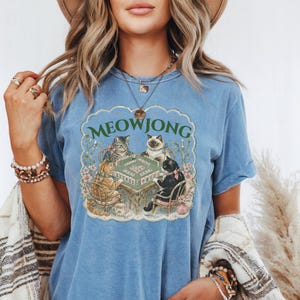 K&ouml;nnte beinhalten: Ein blaues T-Shirt im Jeans-Look mit einem Grafikdruck von vier Katzen, die an einem Tisch sitzen und ein Brettspiel spielen. Der Text MEOWJONG ist in Gr&uuml;n &uuml;ber den Katzen aufgedruckt. Das T-Shirt hat kurze &Auml;rmel und einen Rundhalsausschnitt.