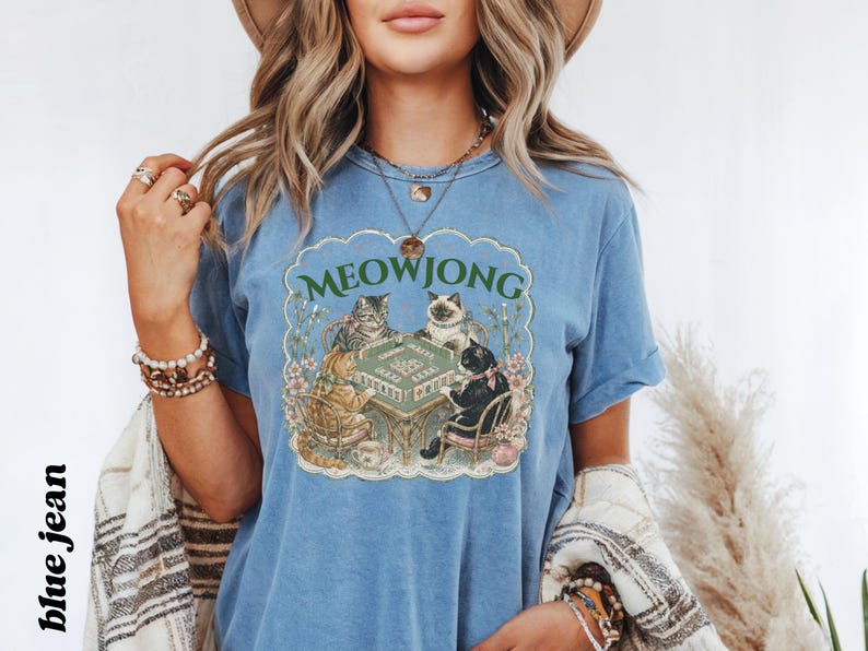 K&ouml;nnte beinhalten: Ein blaues T-Shirt im Jeans-Look mit einem Grafikdruck von vier Katzen, die an einem Tisch sitzen und ein Brettspiel spielen. Der Text MEOWJONG ist in Gr&uuml;n &uuml;ber den Katzen aufgedruckt. Das T-Shirt hat kurze &Auml;rmel und einen Rundhalsausschnitt.