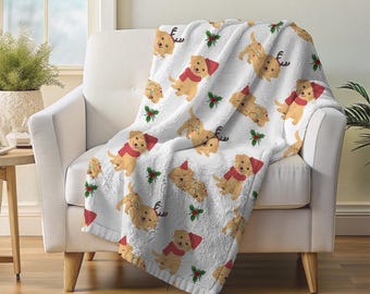 Könnte beinhalten: Eine weiße Fleece-Decke mit einem sich wiederholenden Muster von Golden Retriever-Welpen, die Weihnachtsmützen, Rentiergeweihe und Lichterketten tragen. Die Decke ist über einem weißen Sessel drapiert, mit Stechpalmenzweigen verstreut.
